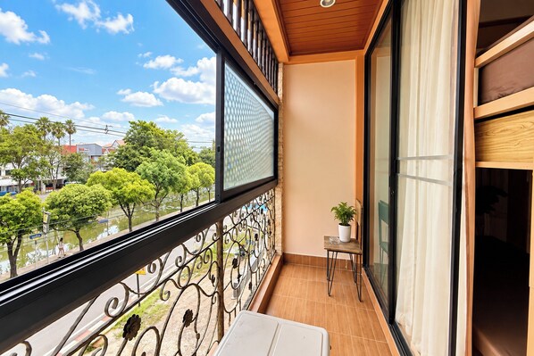 Un balcón con vistas a árboles y edificios, una pequeña mesa con una planta y una ventana con barandilla de metal.