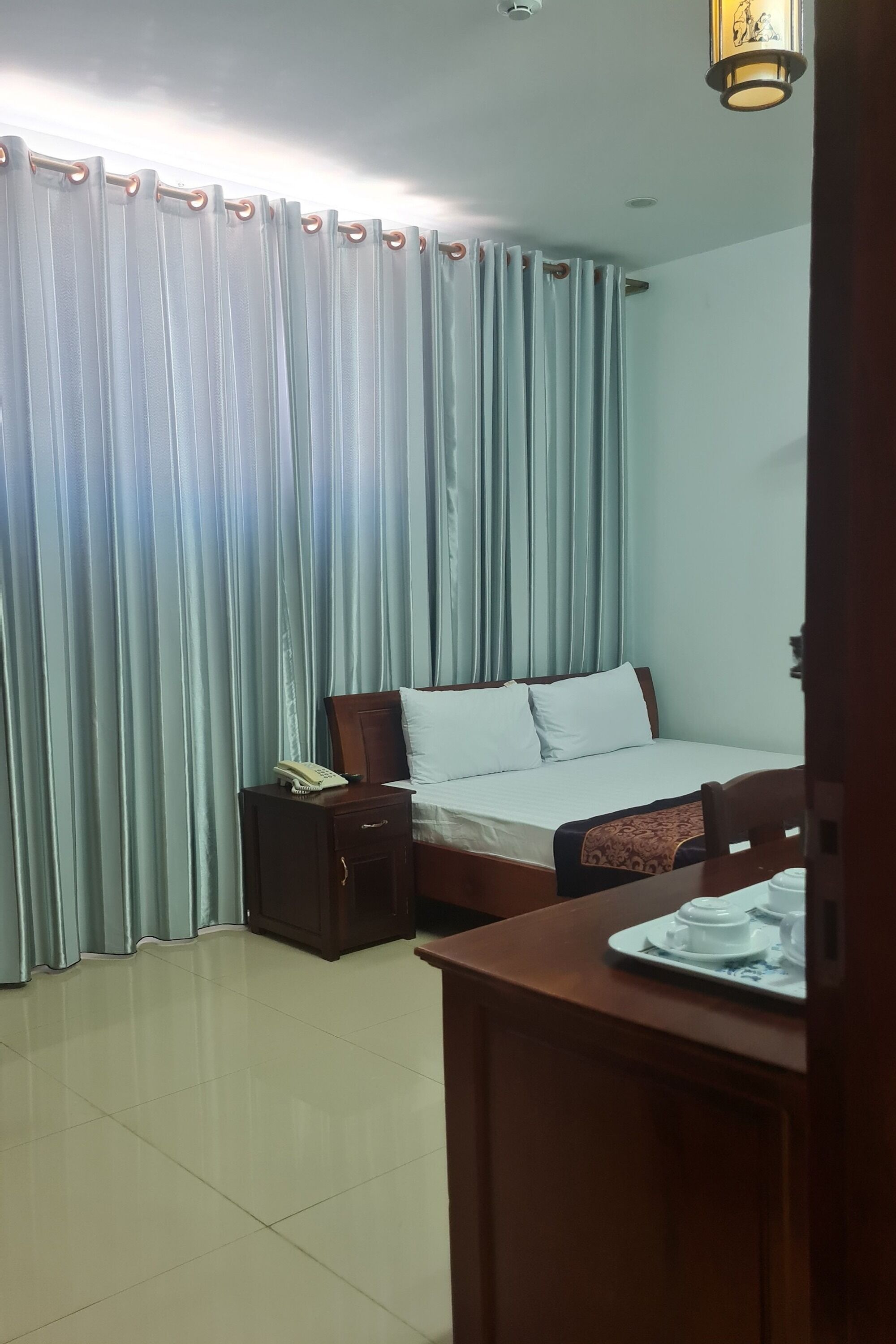 A hotel room with a bed, a bedside table with a phone, a small cabinet, and a table with a teapot and cup.