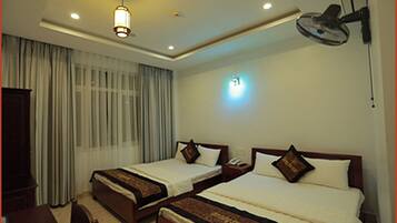 Une chambre d’hôtel avec deux lits simples, un bureau, une chaise, une lampe de bureau, un ventilateur de plafond et une fenêtre avec des rideaux.