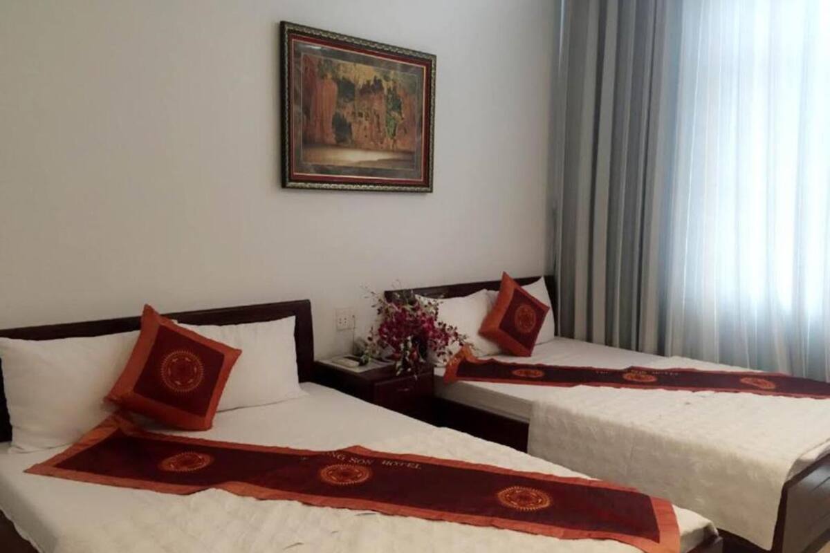 Ein Hotelzimmer mit zwei Betten, beide mit rotem Teppichläufer und weißer Bettwäsche. Darüber an der Wand hängt ein Gemälde.