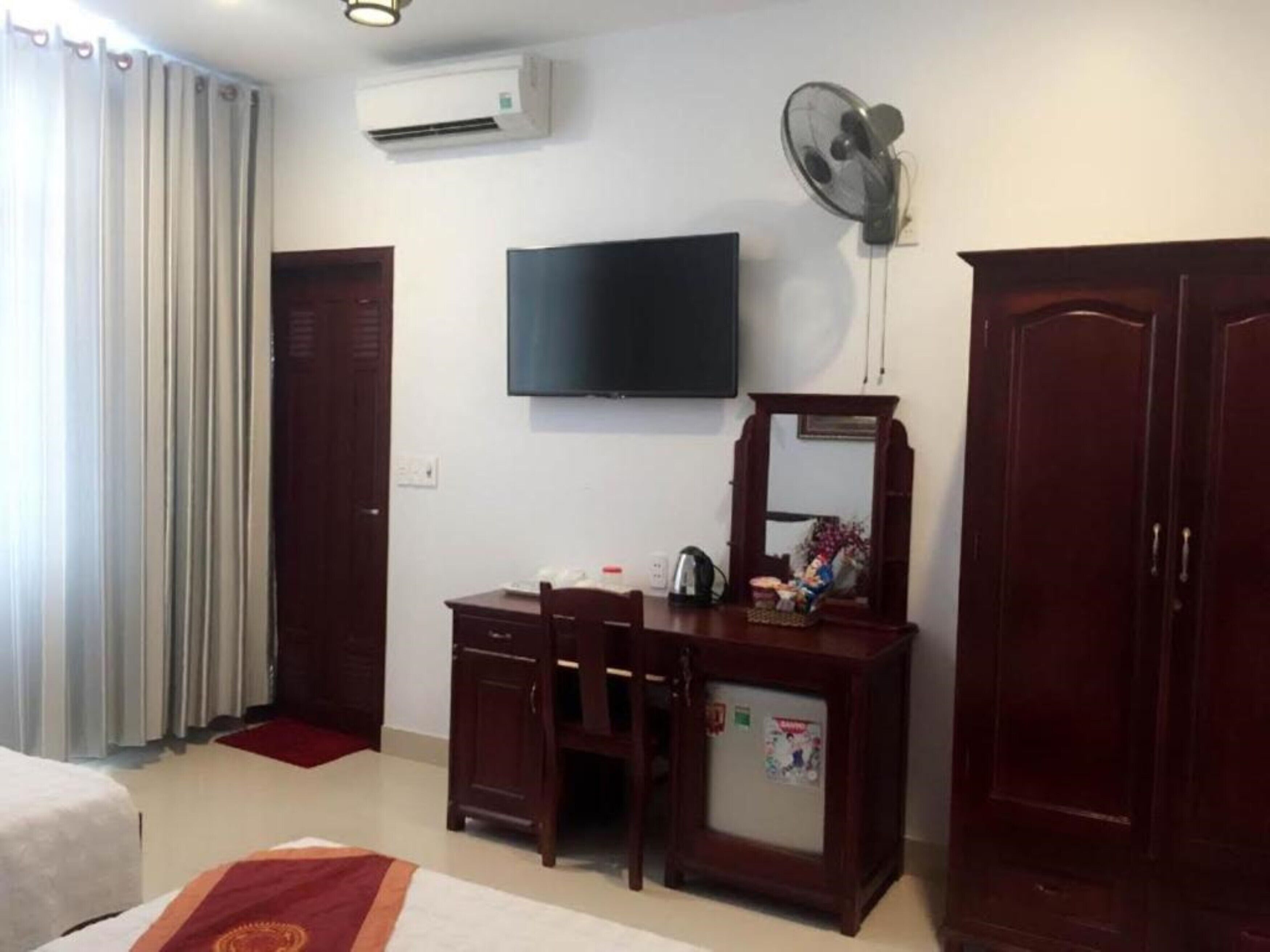 A hotel room with a bed, a desk with a chair, a television, a wardrobe, and an air conditioning unit.