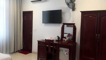 A hotel room with a bed, a desk with a chair, a television, a wardrobe, and an air conditioning unit.
