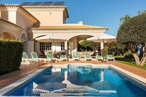 A swimming pool with lounge chairs and umbrellas, surrounded by a building with a tiled roof and a pergola.