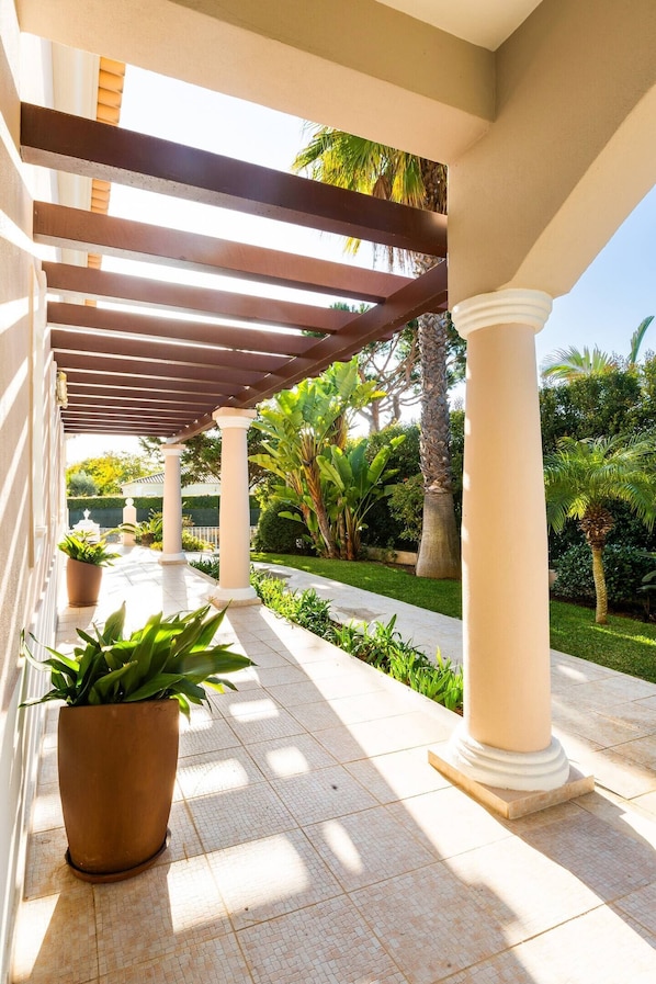 A covered walkway with columns, potted plants, and a garden with palm trees.