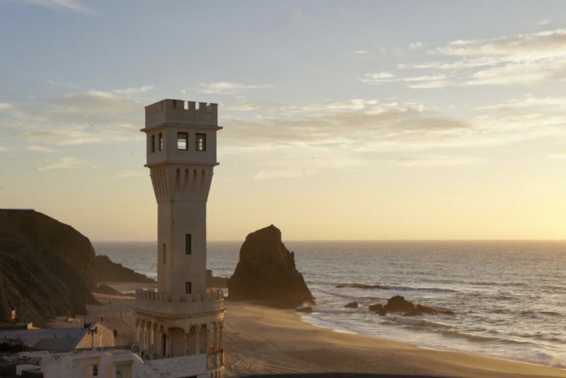 Un phare côtier doté d’une tour et d’une base, surplombant une plage parsemée de rochers et donnant sur la mer.