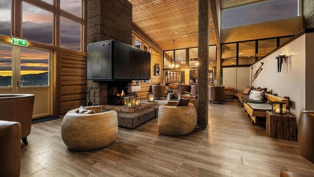 A cozy lounge area with a fireplace, comfortable seating, and large windows offering a view of the sunset. - Spåtind Fjellhotell (Nordre Land)