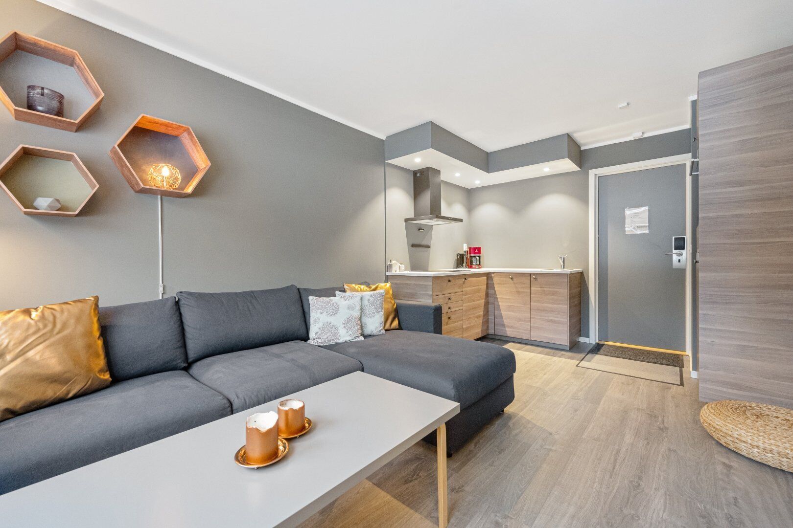 Una sala moderna con un sofá gris, una mesa de centro blanca y una cocina con armarios de madera y horno empotrado.