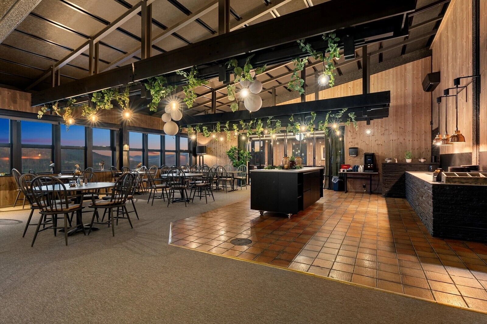 Un ristorante con soffitto alto, travi a vista e un'isola centrale. I tavoli sono disposti attorno all'isola e l'ambiente è ben illuminato da lampade a sospensione.