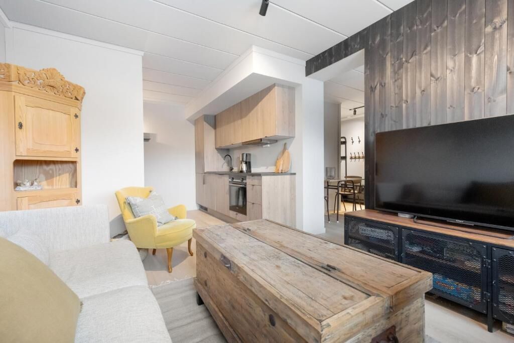 A modern living room with a sofa, yellow armchair, wooden coffee table, TV, and kitchen area.