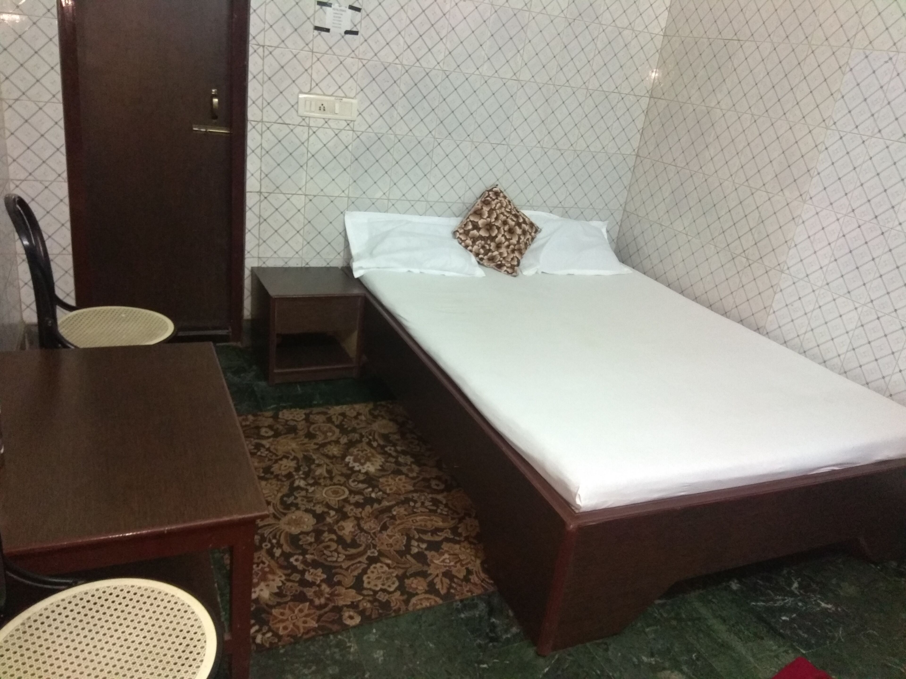 A small, neatly arranged room with a single bed, a wooden nightstand, a chair, and a small table.