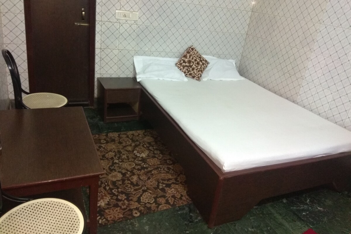 A small, neatly arranged room with a single bed, a wooden nightstand, a chair, and a small table.