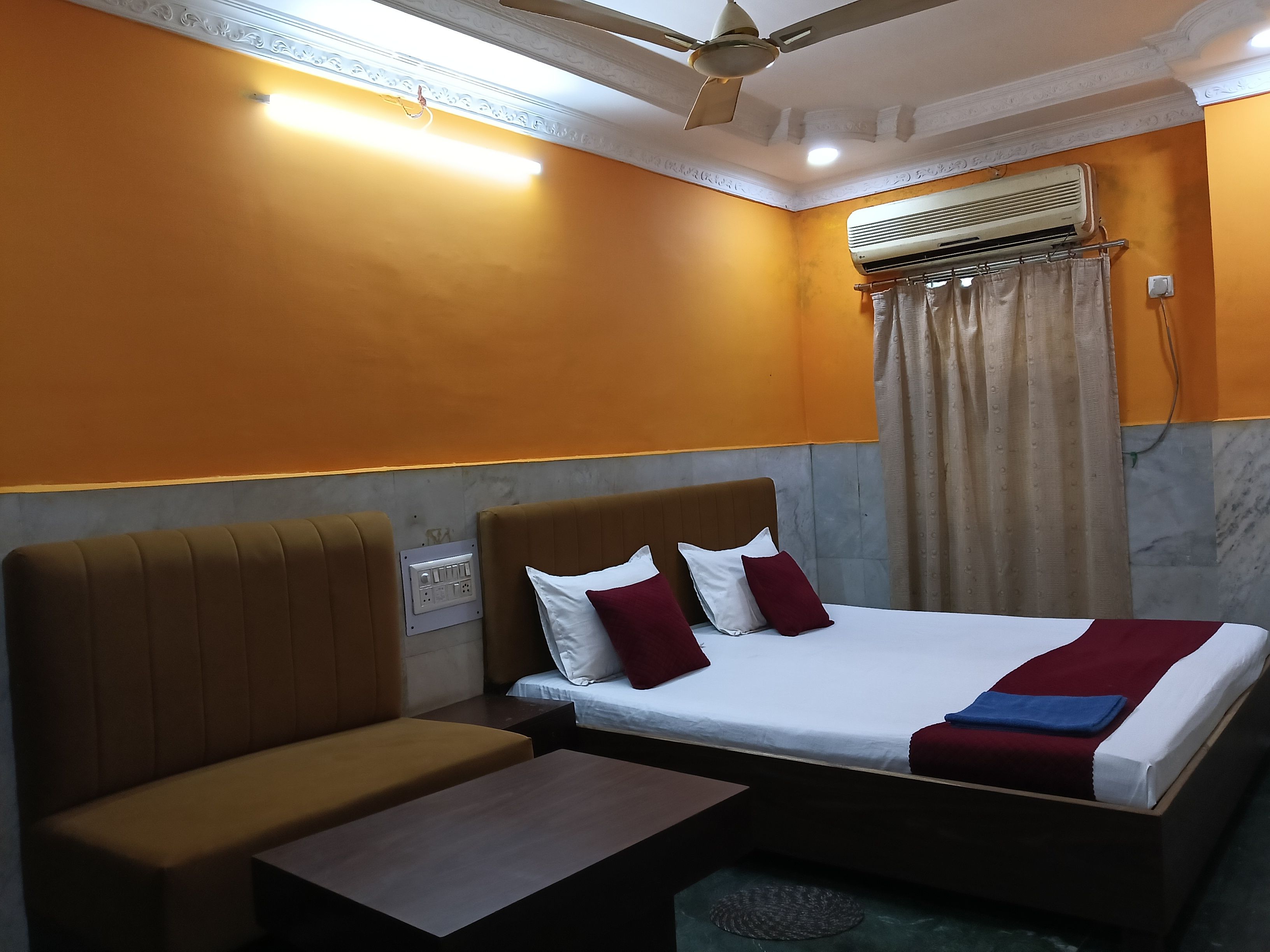 A hotel room with a bed, a sofa, a small table, an air conditioning unit, and a ceiling fan.
