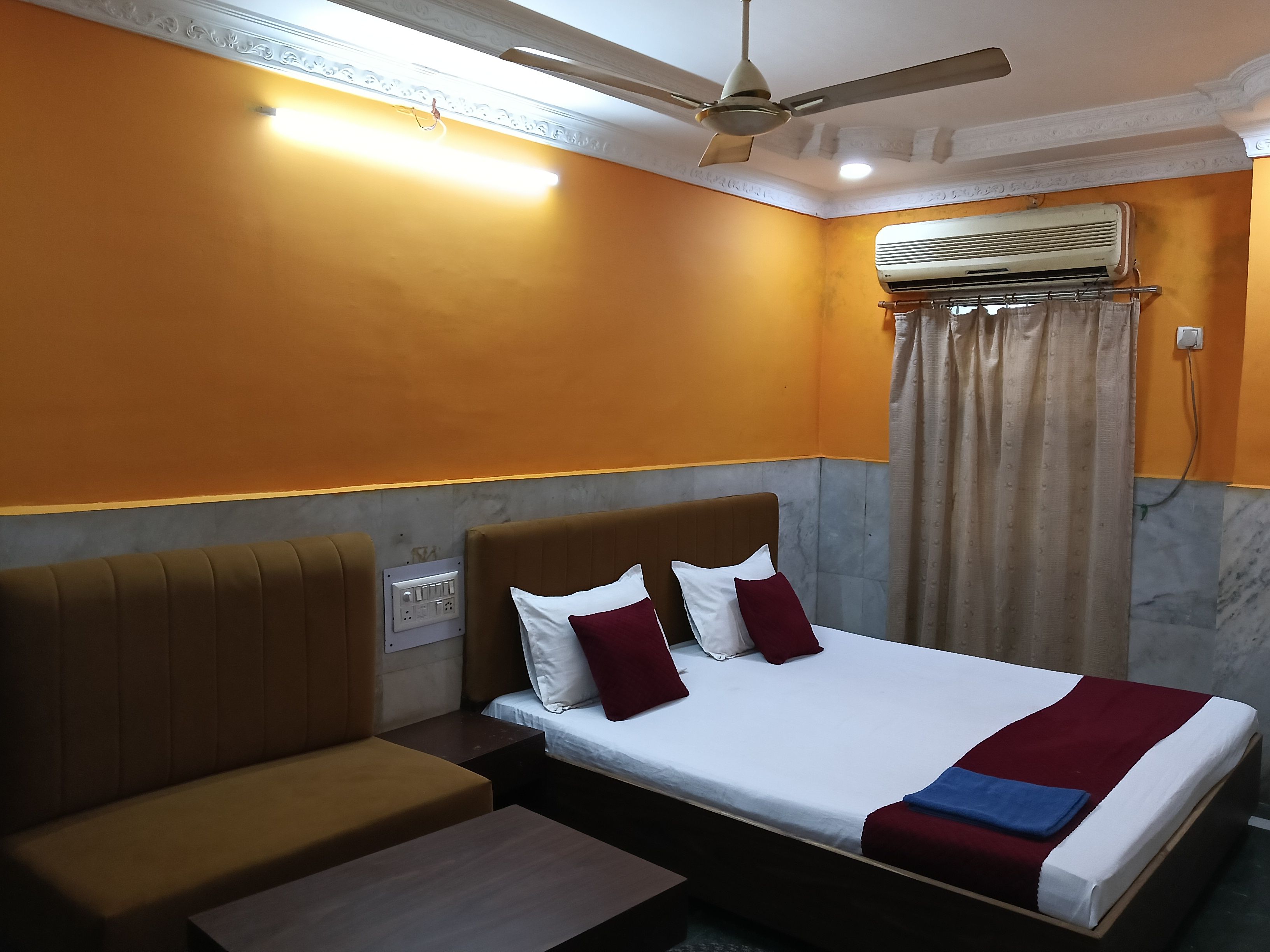 A hotel room with a bed, a sofa, a ceiling fan, an air conditioning unit, and a power outlet.