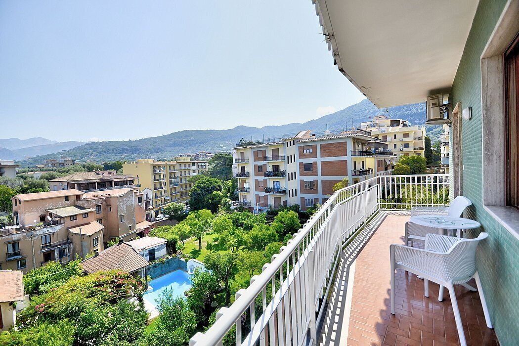 Un balcone con mobili in rattan bianco, vista su una zona residenziale con edifici e una montagna sullo sfondo.