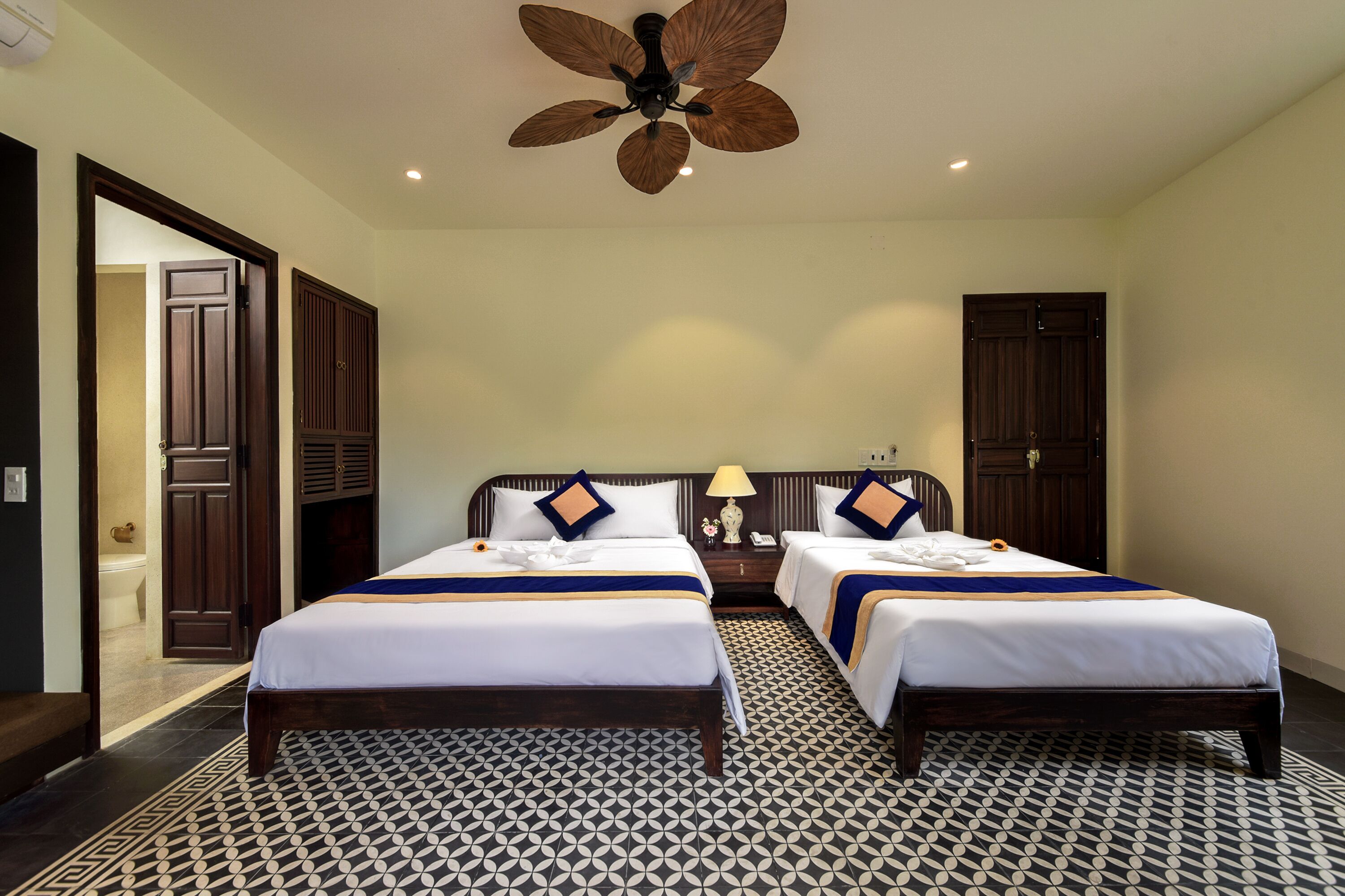 Een hotelkamer met twee bedden, een plafondventilator, een vloer met een patroon en houten deuren.