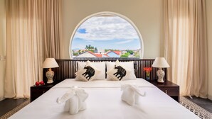 Hotelkamer met een groot bed, twee kussens met olifantmotieven, uitzicht op een stadsbeeld door een groot raam, en een nachtkastje met een lamp en een bloemenvaas.