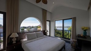 Een hotelkamer met een groot bed, een balkon met uitzicht op een meer en gebouwen, en een plafondventilator.