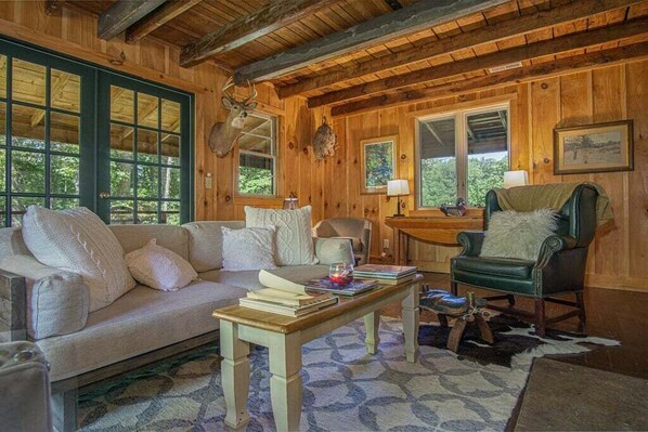 A cozy cabin interior with a wooden ceiling, a sofa, a chair, a table, and a deer head mounted on the wall.