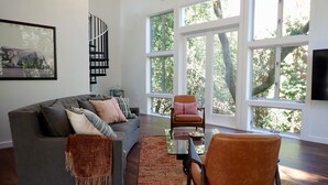 A modern living room with a grey sofa, a brown armchair, and a glass coffee table. There are large windows with a view of trees. - Gorgeous 2Bd + Loft with Deck and Gardens. Soaring 20 ft ceilings, huge windows! (Kentfield)