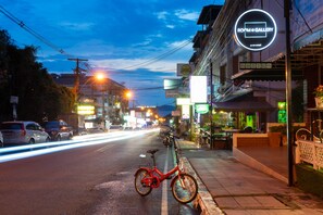 A street view at dusk with a bicycle parked on the sidewalk, buildings with illuminated signs, and a clear sky.