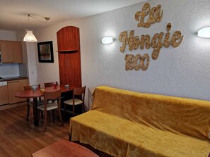 Una sala de estar acogedora con un sofá amarillo, una mesa de comedor de madera con cuatro sillas y una cocina al fondo.
