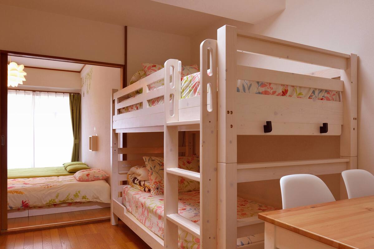 A bunk bed room with a desk and chairs, wooden flooring, and a window with curtains.