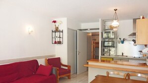 Un ambiente compatto con un divano rosso, un tavolo da pranzo in legno e sedie, un angolo cottura con fornelli e forno, e una piccola panca in legno.