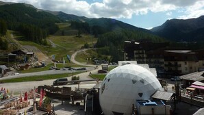 Paesaggio montuoso con una grande struttura a cupola bianca, una zona pavimentata con tavoli e sedie, e edifici sullo sfondo.