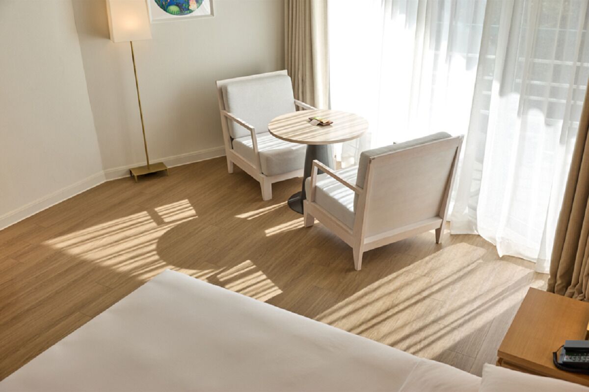 A hotel room with a bed, two white armchairs, a small round table, and a lamp.