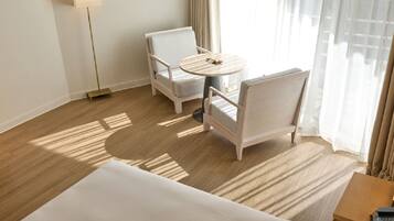 Una habitación de hotel con una cama, dos sillones blancos, una mesita redonda y una lámpara.