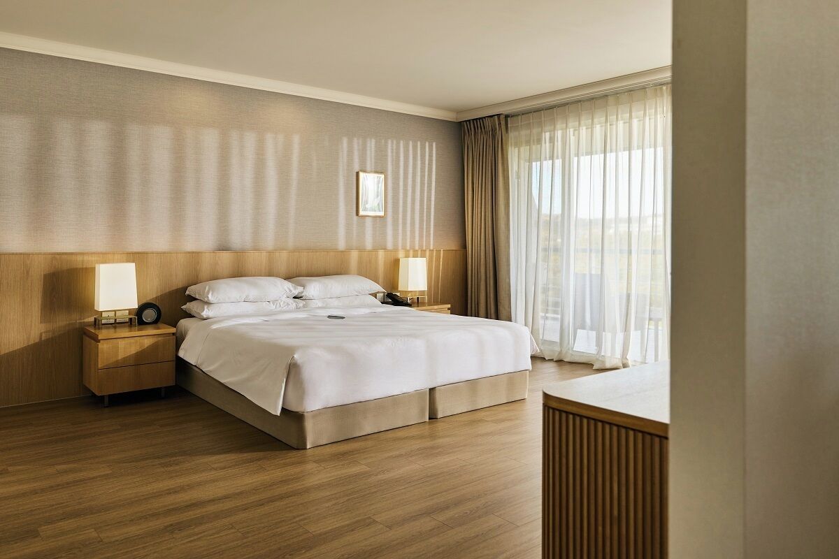 A hotel room with a large bed, wooden headboard, two bedside tables with lamps, a bedside clock, and a view of the outdoors through sheer curtains.