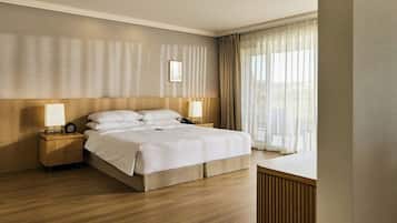 A hotel room with a large bed, wooden headboard, two bedside tables with lamps, a bedside clock, and a view of the outdoors through sheer curtains.