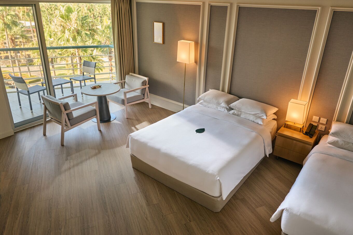 A hotel room with a large bed, a desk with a chair, a small table, and a balcony with chairs.