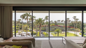 A modern hotel room with a large window offering a view of a landscaped garden with palm trees and a red pathway. - Grand Josun Jeju (Seogwipo)