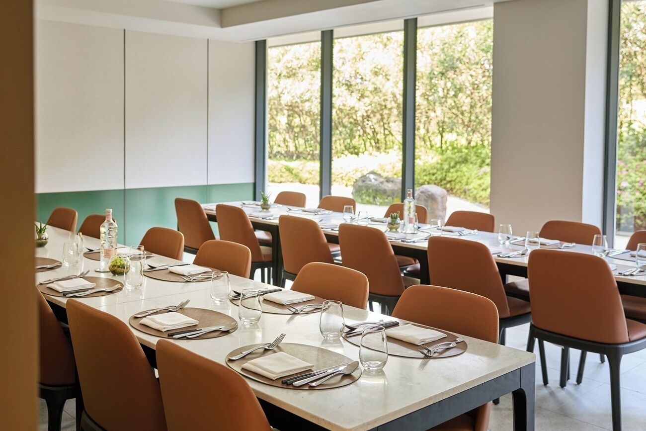A dining area with multiple tables set for meals, large windows with a view of greenery, and a modern interior design.