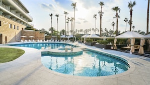 A swimming pool with lounge chairs and umbrellas, surrounded by palm trees and a building with balconies. - Grand Josun Jeju (Seogwipo)