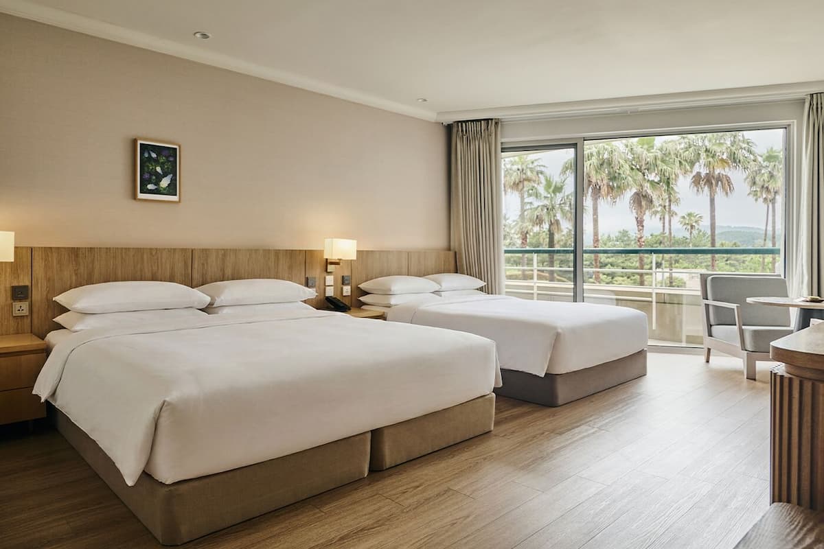 Habitación de hotel con dos camas, un escritorio, una silla y un ventanal con vista a palmeras.