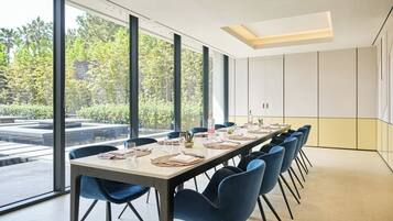 A conference room with a long table, blue chairs, and large windows offering a view of greenery outside.