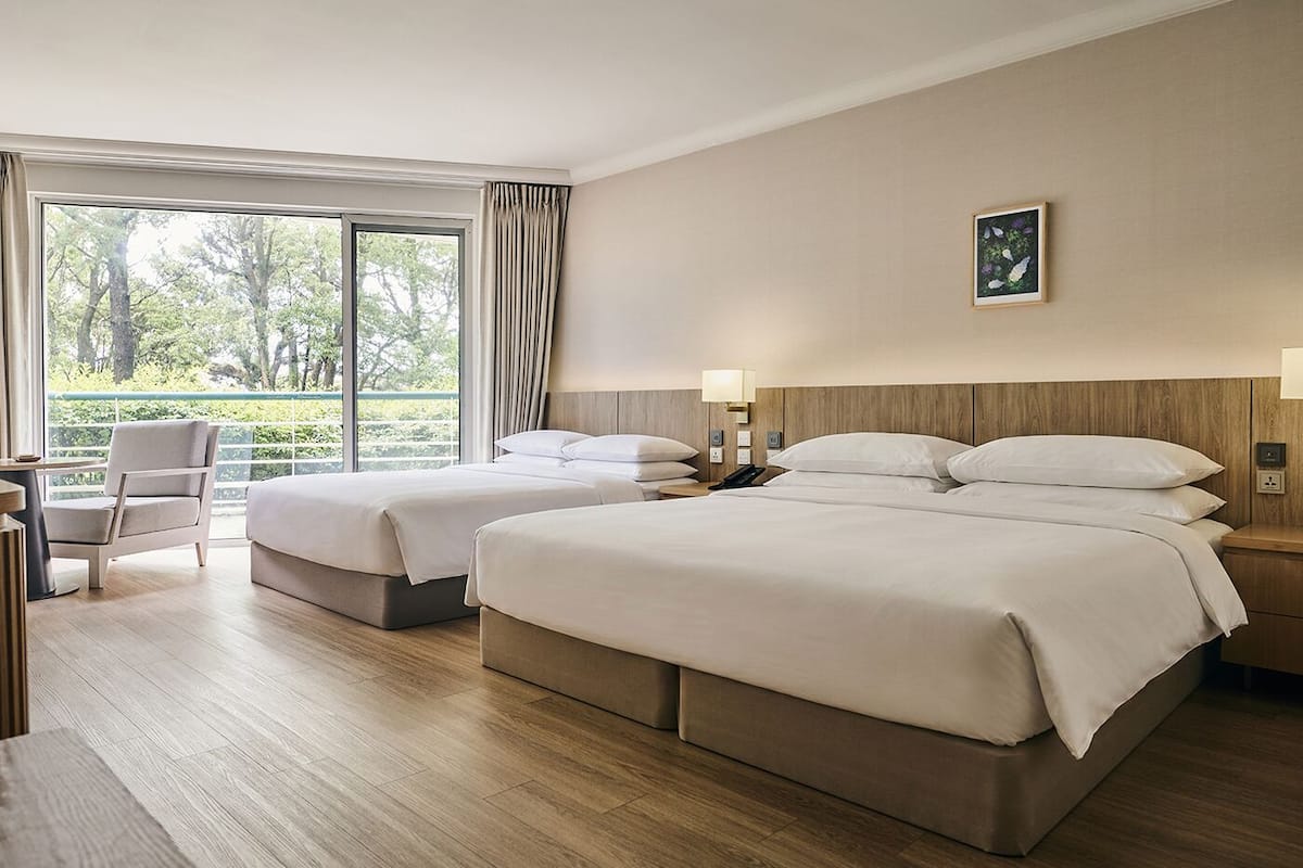 Habitación de hotel con dos camas, un escritorio, una silla y un ventanal con cortinas.