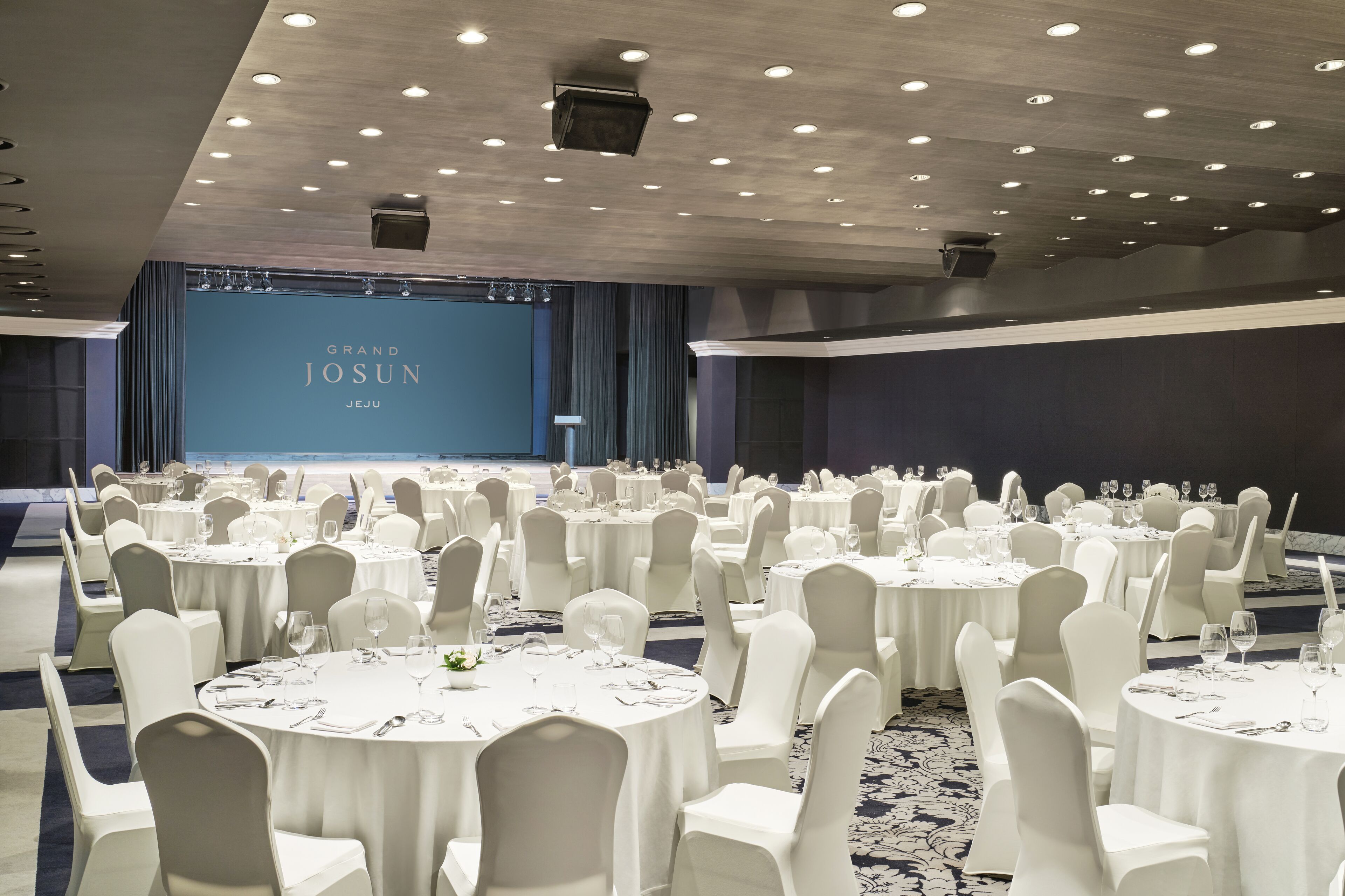 A banquet hall with round tables set for a formal event, a stage with a screen, and a dark blue curtain backdrop.