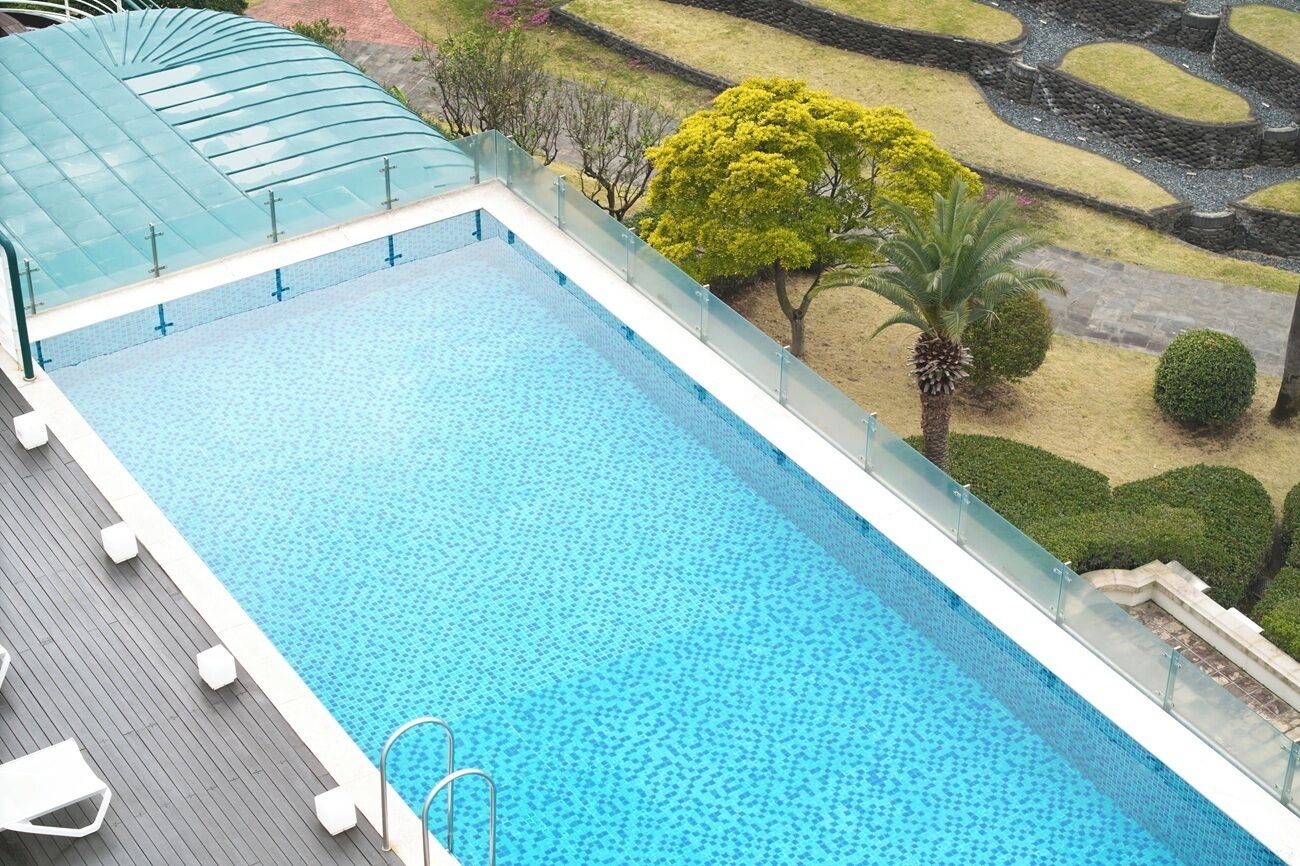 A rectangular swimming pool with a glass enclosure, surrounded by a deck with white lounge chairs and a small staircase leading to a covered area.