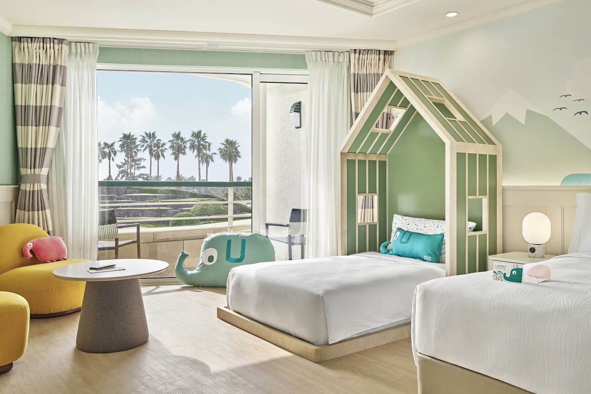 Habitación de hotel con dos camas, una mesita, una silla y vistas a palmeras.