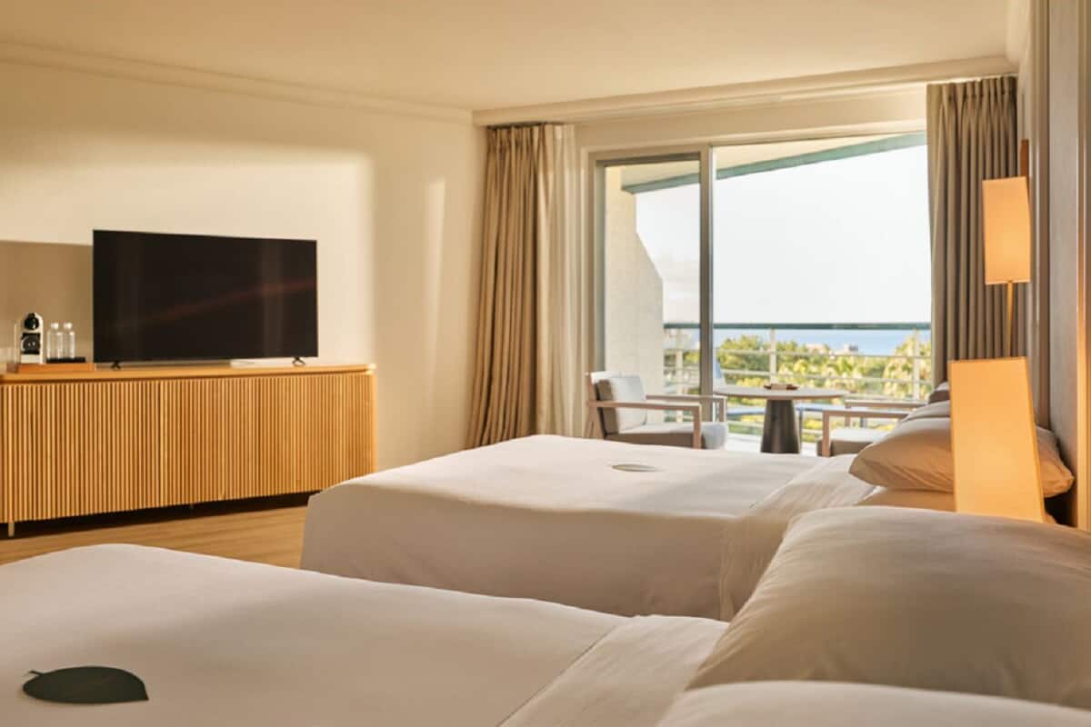 Habitación de hotel con dos camas, un televisor en un mueble de madera y un balcón con vista al mar.