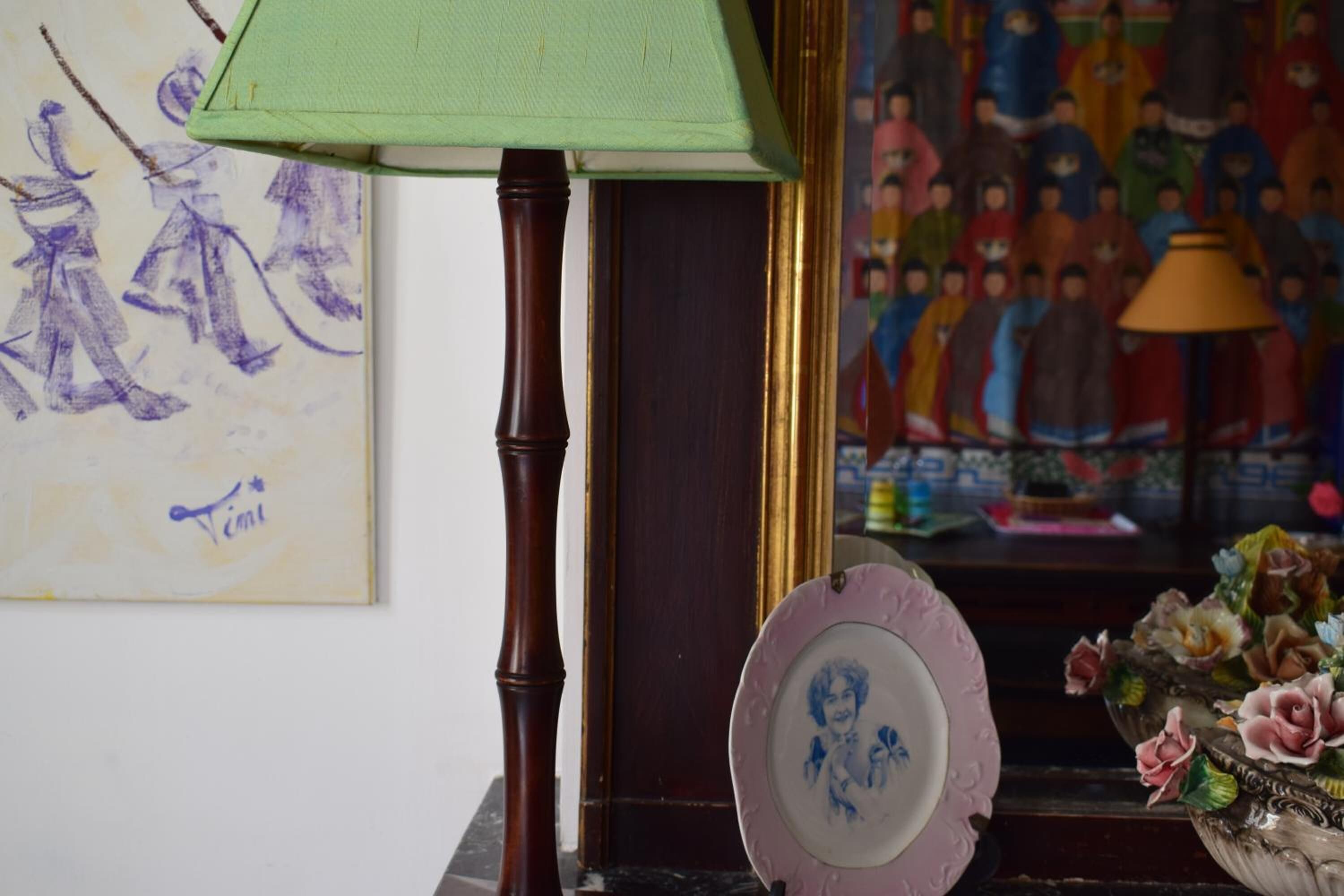 Une pièce avec une lampe verte, une œuvre d’art encadrée, une assiette décorative et un vase rempli de fleurs.