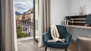 A modern room with a large window, a blue armchair, a small table, and a view of a cityscape. - Elys Suites (Sorrento)