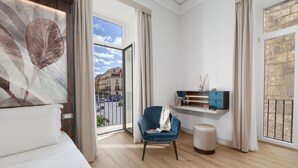 A modern hotel room with a large window, a blue chair, a bedside table with a lamp, and a wall-mounted shelf. - Elys Suites (Sorrento)