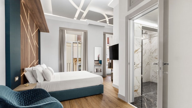 A modern bedroom with a bed, a blue chair, a TV, and a bathroom visible through an open door. - Elys Suites (Sorrento)