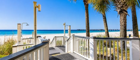 A wooden deck leading to a beach with palm trees, a pier in the distance, and clear blue skies.