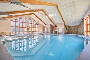 An indoor swimming pool with clear glass walls, wooden beams, and a stone wall. - Résidence Odalys Rochebrune Les Cimes (Orcieres)