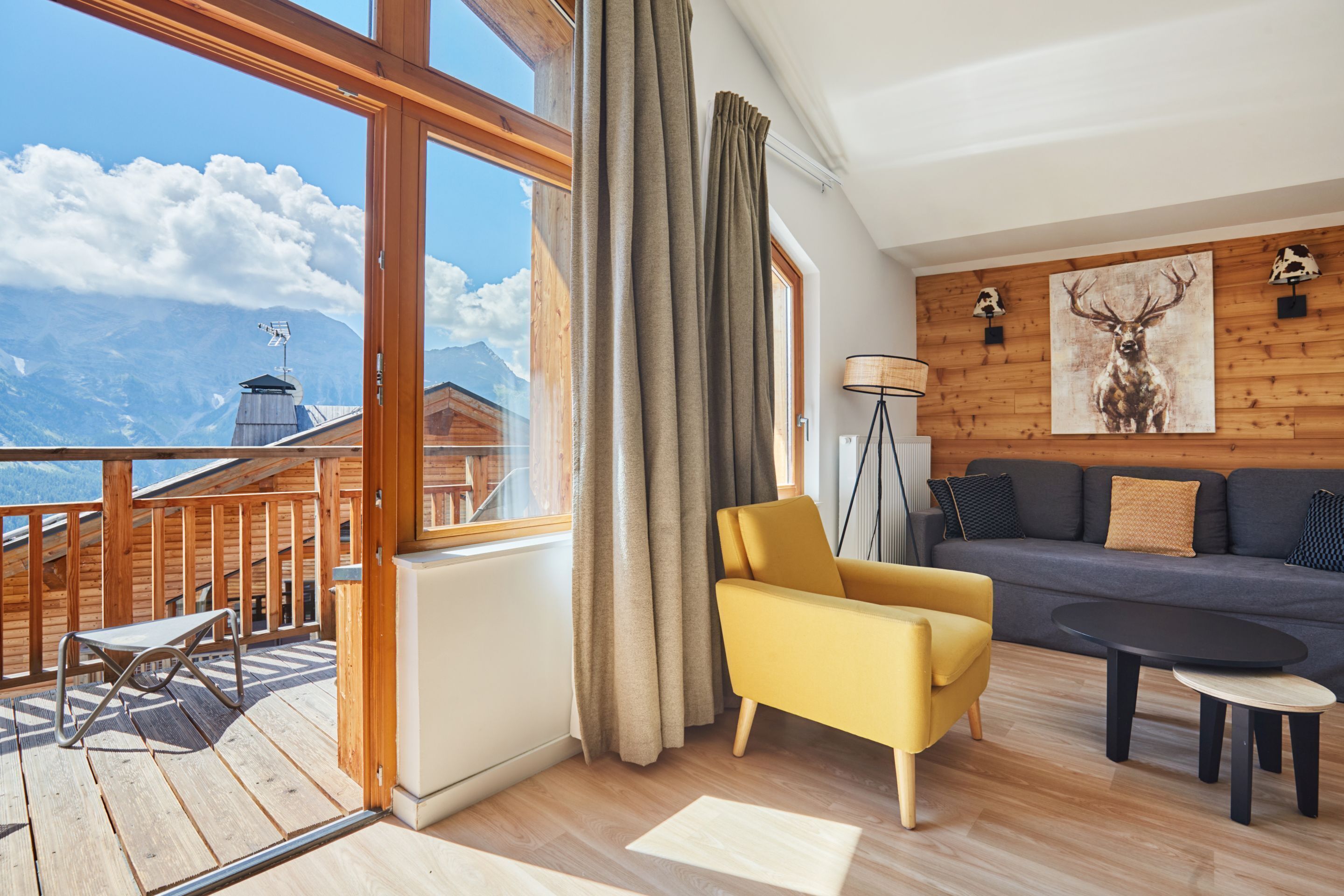 Ein modernes Wohnzimmer mit einer Couch, einem gelben Sessel und einem Couchtisch. Ein großes Fenster bietet Blick auf Berge und eine Holzterrasse.